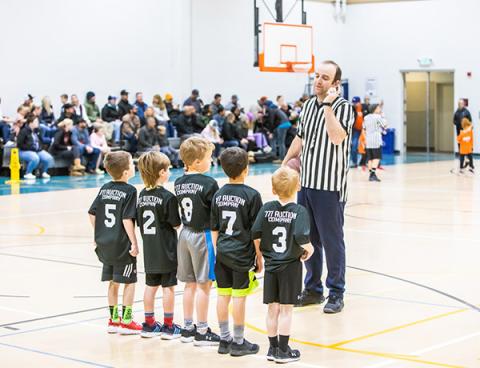 Kindergarten age kids in basketball uniforms getting direction from the referee.