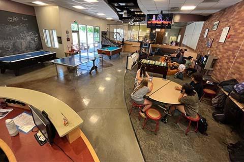 Image of Teen Center space, with youth sitting at a round table and a variety of game options, such as pool and table tennis in the background.