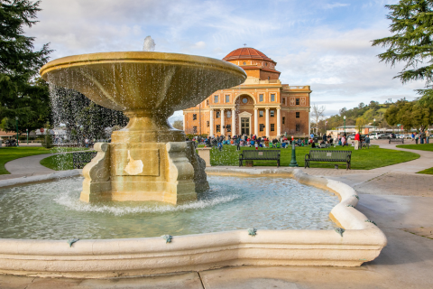 Large fountain in the foreground with a crowd of people on a stretch of grass in the Sunken Gardens park with the Historic Atascadero City Hall building in the background.